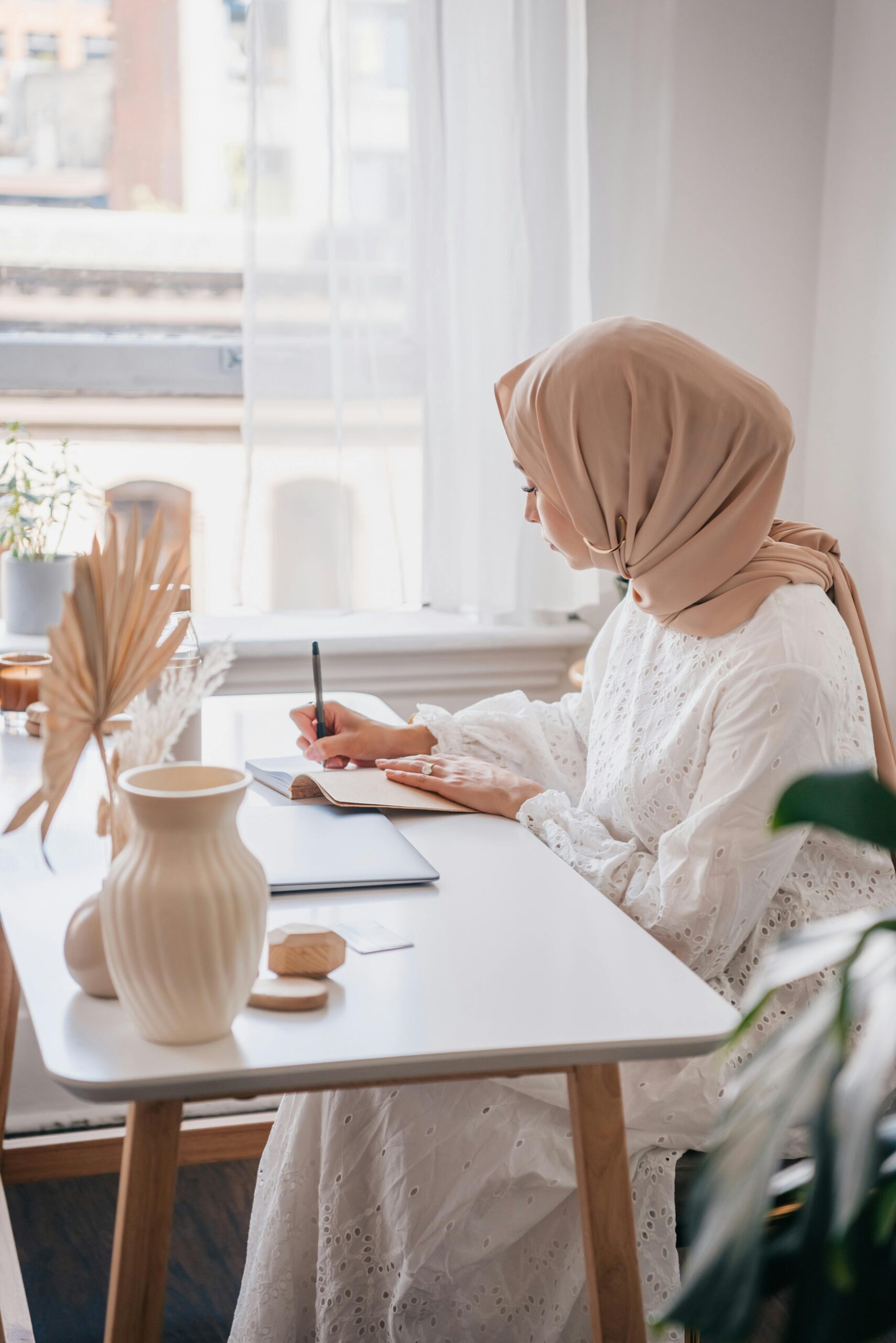 A woman in a peach hijab writes in a notebook at her desk by a window